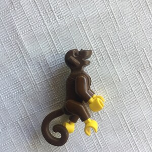 May include: A brown plastic monkey figurine with yellow hands and feet. The monkey is sitting with its tail curled behind it.