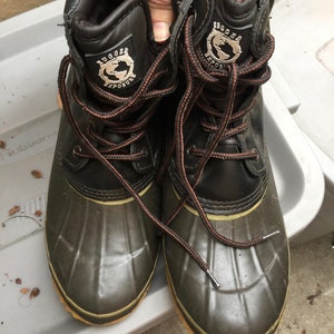 May include: A pair of black and green leather winter boots with brown laces. The boots have a logo that says "Rugged Exposure" with a globe graphic.