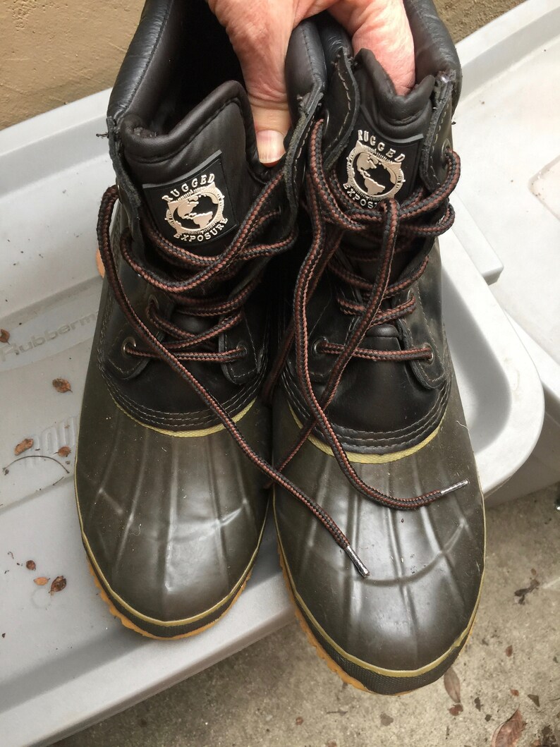 May include: A pair of black and green leather winter boots with brown laces. The boots have a logo that says "Rugged Exposure" with a globe graphic.