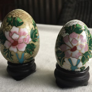 May include: Two decorative eggs with floral designs, painted in shades of green, white, pink, and gold. The eggs are on small black wooden stands.