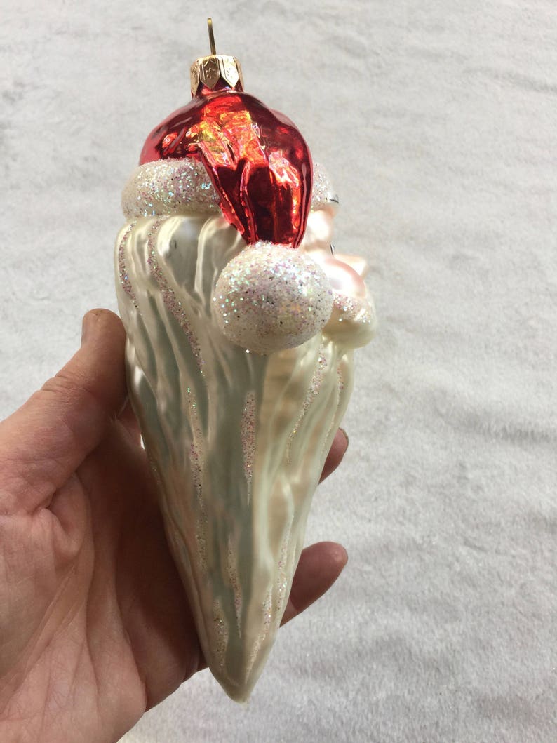 以下が含まれることがあります： サンタクロースの形をしたクリスマスオーナメント。オーナメントは、白いグリッタートリムが付いた赤い帽子、白いひげ、真珠色のボディが特徴です。オーナメントは手で持たれています。