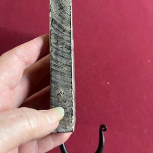 May include: A handcrafted wooden wall hook with a dark, curved metal hook. The wood has a natural, weathered appearance with visible grain and a light gray and white color. The hook is attached to the bottom of the wooden block.