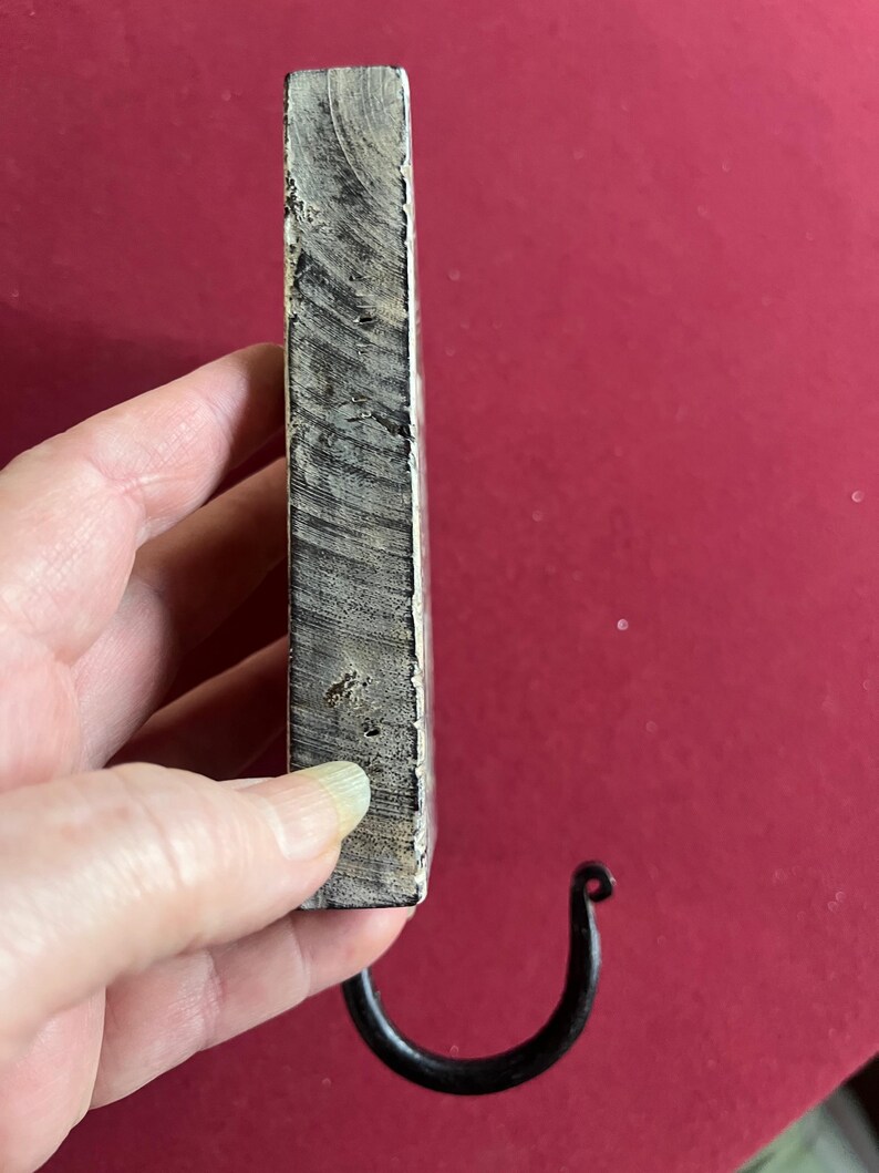 May include: A handcrafted wooden wall hook with a dark, curved metal hook. The wood has a natural, weathered appearance with visible grain and a light gray and white color. The hook is attached to the bottom of the wooden block.