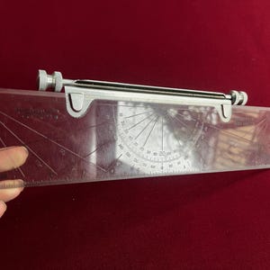 Könnte beinhalten: Ein transparenter Kunststoff-Winkelmesser mit einem Metalllineal und Einstellknöpfen. Der Winkelmesser hat Gradmarkierungen und ein Lineal entlang einer Kante. Das Werkzeug dient zum Messen und Zeichnen von Winkeln.