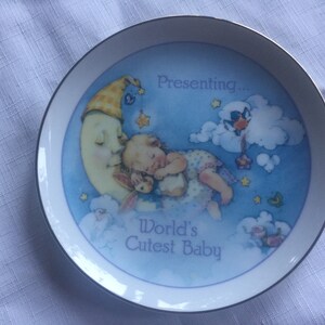 Pode incluir: Um prato de cer&acirc;mica branco com fundo azul e uma lua crescente com um beb&ecirc; dormindo sobre ela. O prato diz "Presenting... World's Cutest Baby".