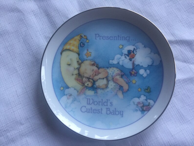 Pode incluir: Um prato de cer&acirc;mica branco com fundo azul e uma lua crescente com um beb&ecirc; dormindo sobre ela. O prato diz "Presenting... World's Cutest Baby".
