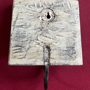 May include: A square, wooden wall hook with a distressed, off-white finish. The hook is made of metal and is attached to the wooden base with screws. A keyhole is visible on the back for hanging.