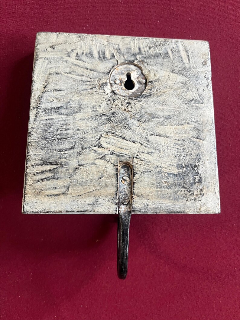 May include: A square, wooden wall hook with a distressed, off-white finish. The hook is made of metal and is attached to the wooden base with screws. A keyhole is visible on the back for hanging.