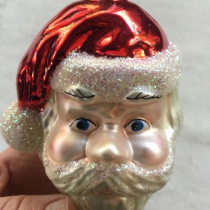 以下が含まれることがあります： サンタクロースの頭の形をしたクリスマスオーナメント。キラキラ光る赤い帽子、青い目、白いひげと口ひげにキラキラのアクセントが付いています。オーナメントは手で持たれています。