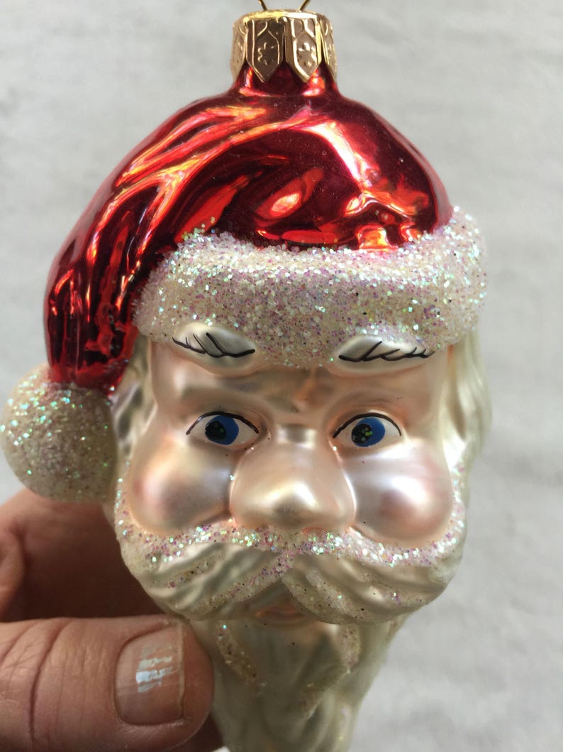 以下が含まれることがあります： サンタクロースの頭の形をしたクリスマスオーナメント。キラキラ光る赤い帽子、青い目、白いひげと口ひげにキラキラのアクセントが付いています。オーナメントは手で持たれています。