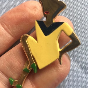 Puede incluir: Un broche dorado que representa a una mujer estilizada con un vestido amarillo y un cuello negro y azul. La mujer est&aacute; sentada con las piernas cruzadas y lleva zapatos verdes.