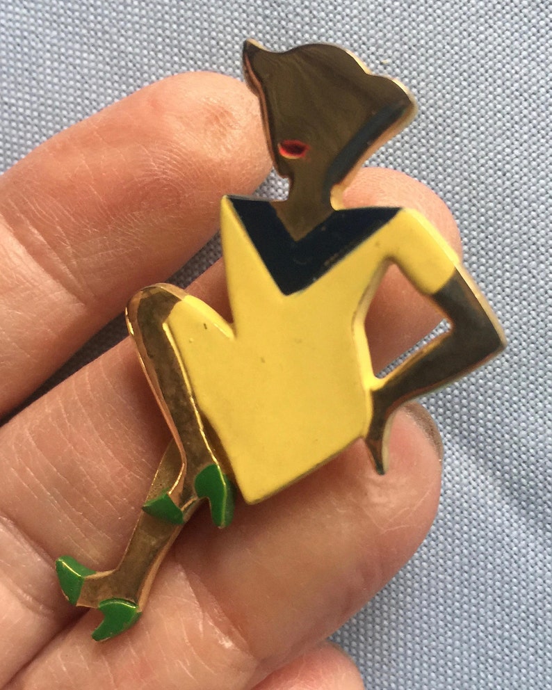 Puede incluir: Un broche dorado que representa a una mujer estilizada con un vestido amarillo y un cuello negro y azul. La mujer est&aacute; sentada con las piernas cruzadas y lleva zapatos verdes.