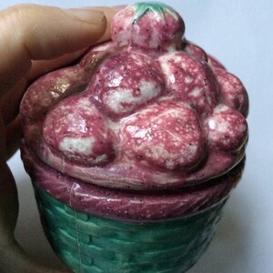 Può includere: Un vaso di ceramica a forma di cesto pieno di fragole rosa e bianche. Anche il coperchio è a forma di fragola.