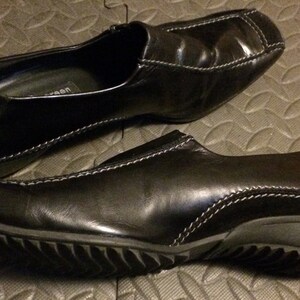Puede incluir: Un par de zapatos negros de cuero sin cordones con un detalle de costura blanca. Los zapatos tienen una suela de goma texturizada.