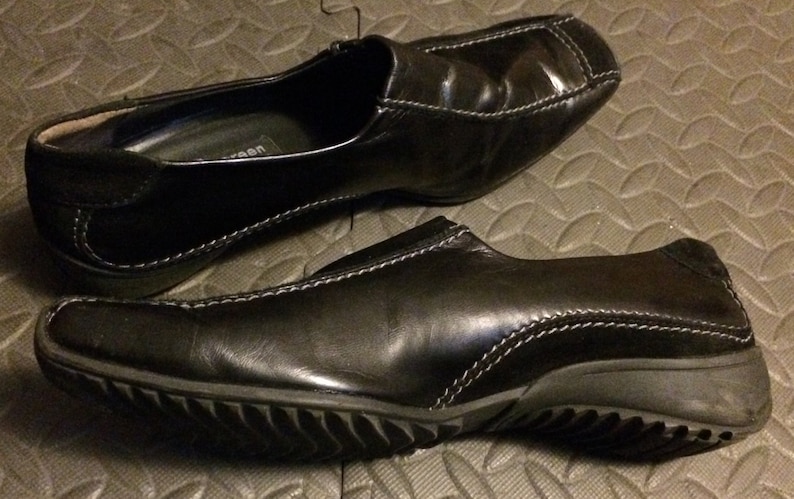 Puede incluir: Un par de zapatos negros de cuero sin cordones con un detalle de costura blanca. Los zapatos tienen una suela de goma texturizada.