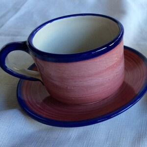 May include: A pink ceramic teacup and saucer with a blue rim. The cup has a white interior and the saucer has a swirling pattern.