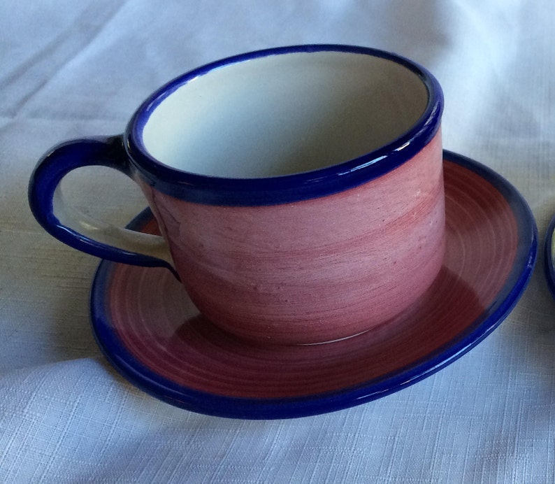 May include: A pink ceramic teacup and saucer with a blue rim. The cup has a white interior and the saucer has a swirling pattern.