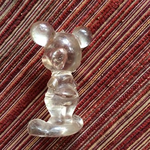 Puede incluir: Una figurita de cristal transparente de Mickey Mouse. La figurita est&aacute; de pie con los brazos a los lados y la cabeza ligeramente inclinada hacia un lado. La figurita est&aacute; sobre un fondo de rayas rojas y blancas.