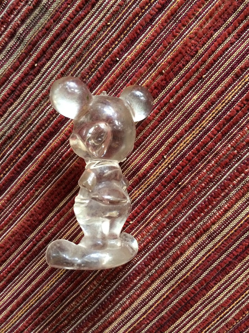 Puede incluir: Una figurita de cristal transparente de Mickey Mouse. La figurita est&aacute; de pie con los brazos a los lados y la cabeza ligeramente inclinada hacia un lado. La figurita est&aacute; sobre un fondo de rayas rojas y blancas.