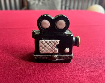 May include: A black ceramic vintage movie camera trinket box with pink details. The box has a hinged lid and a small gold-colored handle on the side.