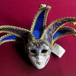 May include: A white and blue jester mask with gold accents and a blue and gold feathered headdress. The mask has a painted design of flowers and leaves. The mask is labeled "Maschera del Carnevale del Venezia".