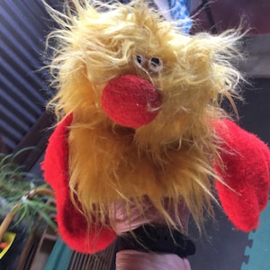 May include: A yellow fuzzy hand puppet with red arms and a red nose. The puppet has two white eyes.
