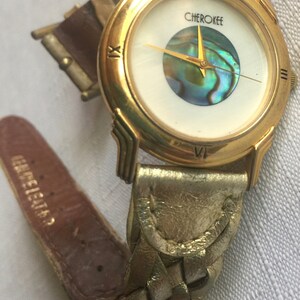 May include: Gold-tone wristwatch with a white face and a mother-of-pearl dial. The watch has a gold-tone band with a braided leather design. The watch face says "Cherokee".