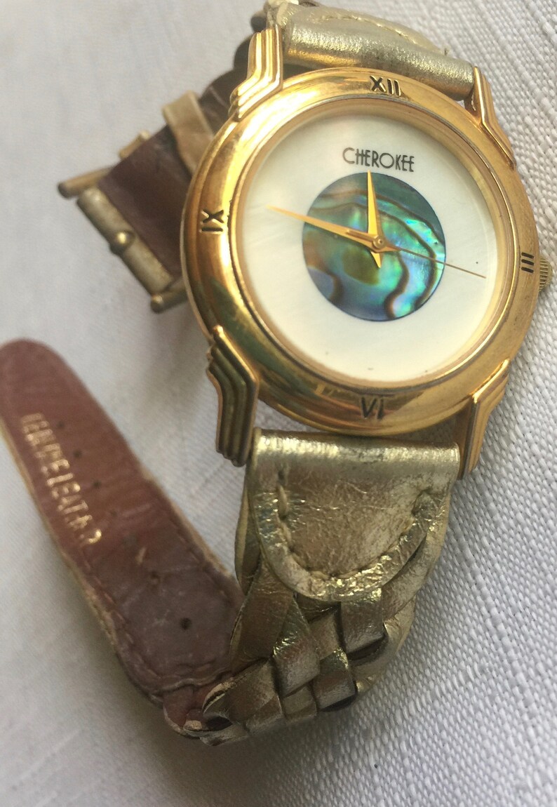 May include: Gold-tone wristwatch with a white face and a mother-of-pearl dial. The watch has a gold-tone band with a braided leather design. The watch face says "Cherokee".