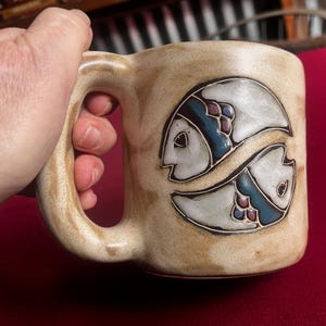 May include: A beige ceramic coffee mug with a brown, blue, and white fish design. The mug has a large handle.