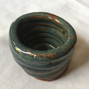 May include: A small, hand-thrown ceramic bowl with a teal glaze and brown accents. The bowl has a rounded shape and a slightly uneven rim.