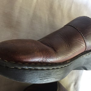 May include: Brown leather clog with a thick black rubber sole. The clog has a stitched seam around the top edge and a strap across the top.