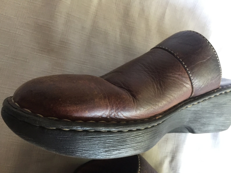 May include: Brown leather clog with a thick black rubber sole. The clog has a stitched seam around the top edge and a strap across the top.