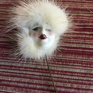 May include: A white fur pin with a porcelain face. The face has blue eyes, red lips, and pink cheeks. The pin is attached to a gold-colored pin back.