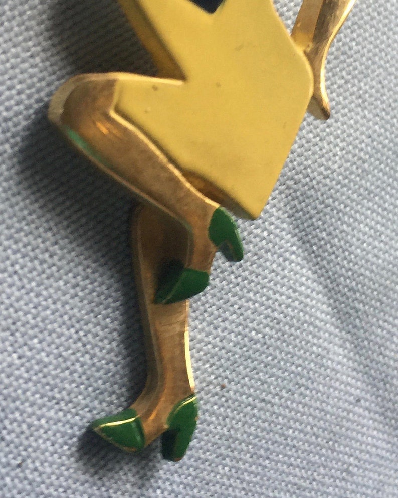Puede incluir: Figurita de metal dorado de una mujer con un vestido amarillo y zapatos verdes. La figurita est&aacute; en posici&oacute;n sentada con una pierna doblada y la otra extendida.