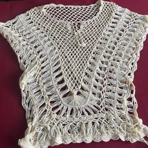 May include: An off-white crochet top featuring an intricate open-work pattern with a V-neckline, short sleeves, and a scalloped hem. The garment is constructed from interconnected yarn loops and knots, creating a textured, semi-sheer design.