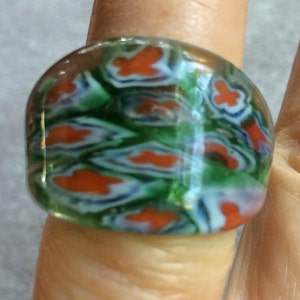 May include: A colorful glass ring with a mosaic pattern. The ring features a mix of orange, green, and blue hues. The design is a repeating pattern of floral-like shapes. The ring is on a finger.