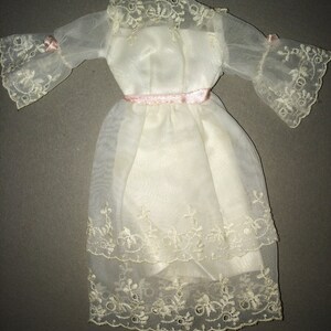 Puede incluir: Un vestido blanco de gasa con delicados detalles de encaje y una cinta de raso rosa alrededor de la cintura.