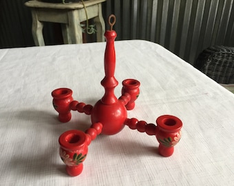 May include: A red wooden candle holder with four candle cups and a central stem. The candle holder has a decorative floral design in green and white. It has a metal loop at the top for hanging.
