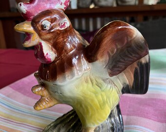 Pode incluir: Uma figura de galo de cerâmica com um design pintado em marrom, amarelo e preto. O galo tem uma crista e barbelas vermelhas e está em pé sobre uma base preta.