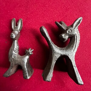 May include: Two small, black cast iron figurines of stylized animals. The animal on the left is a donkey with long ears and a tail. The animal on the right is a unicorn with a single horn and a tail. Both animals are standing on a red surface.