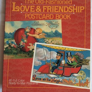 K&ouml;nnte beinhalten: Ein rotes Postkartenbuch mit dem Titel "The Old-Fashioned Love & Friendship Postcard Book". Das Cover zeigt zwei Vintage-Postkarten. Eine zeigt einen Cherub mit einem Herzen, die andere ein Paar in einem fliegenden Fahrzeug. Das Buch enth&auml;lt 40 farbige Postkarten.
