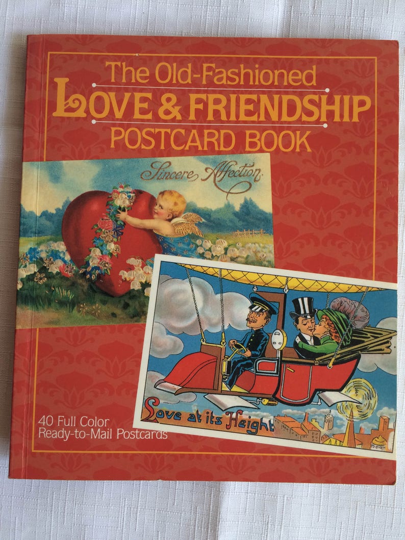 K&ouml;nnte beinhalten: Ein rotes Postkartenbuch mit dem Titel "The Old-Fashioned Love & Friendship Postcard Book". Das Cover zeigt zwei Vintage-Postkarten. Eine zeigt einen Cherub mit einem Herzen, die andere ein Paar in einem fliegenden Fahrzeug. Das Buch enth&auml;lt 40 farbige Postkarten.