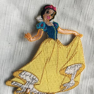May include: Embroidered iron-on patch featuring Snow White from Disney's Snow White and the Seven Dwarfs. The patch shows Snow White in her iconic yellow dress with a blue bodice and red bow.