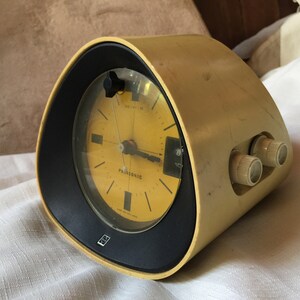 May include: A yellow vintage Panasonic alarm clock with a black face and a round dial. The clock has two knobs on the side for setting the alarm and volume.