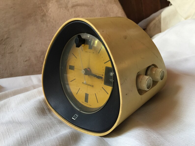 May include: A yellow vintage Panasonic alarm clock with a black face and a round dial. The clock has two knobs on the side for setting the alarm and volume.