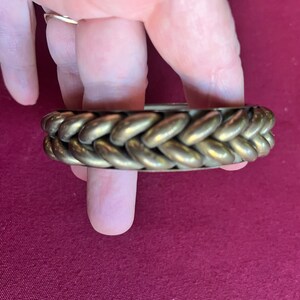 May include: A gold-toned metal bracelet with a braided design. The bracelet is made of a series of connected oval-shaped pieces.