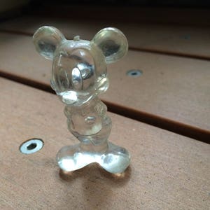 Puede incluir: Una figurita de pl&aacute;stico transparente de Mickey Mouse. La figurita est&aacute; de pie sobre una superficie de madera.