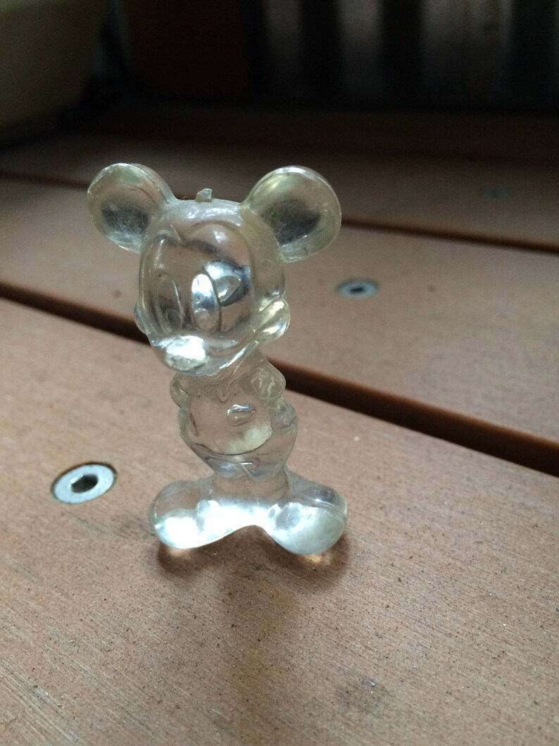 Puede incluir: Una figurita de pl&aacute;stico transparente de Mickey Mouse. La figurita est&aacute; de pie sobre una superficie de madera.