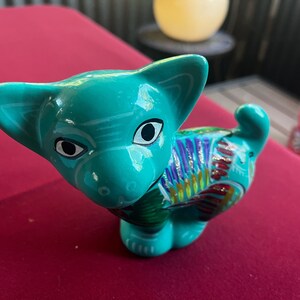 May include: A teal blue ceramic cat figurine with colorful painted details. The cat has large black eyes and a white stripe on its forehead.