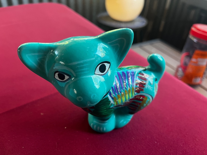 May include: A teal blue ceramic cat figurine with colorful painted details. The cat has large black eyes and a white stripe on its forehead.